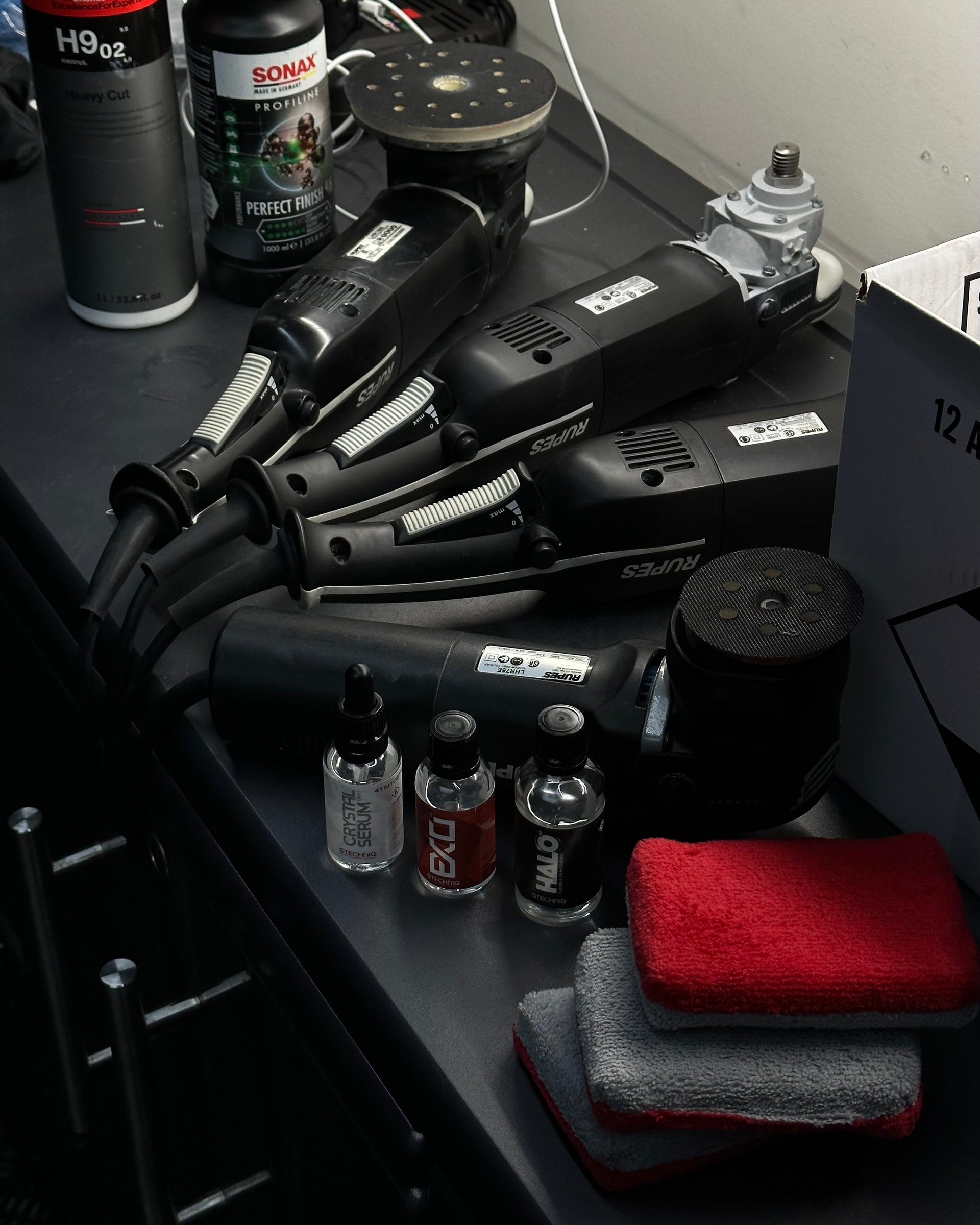 Collection of car detailing tools including polishers, spray bottles, SONAX products, and microfiber cloths organized on a black surface