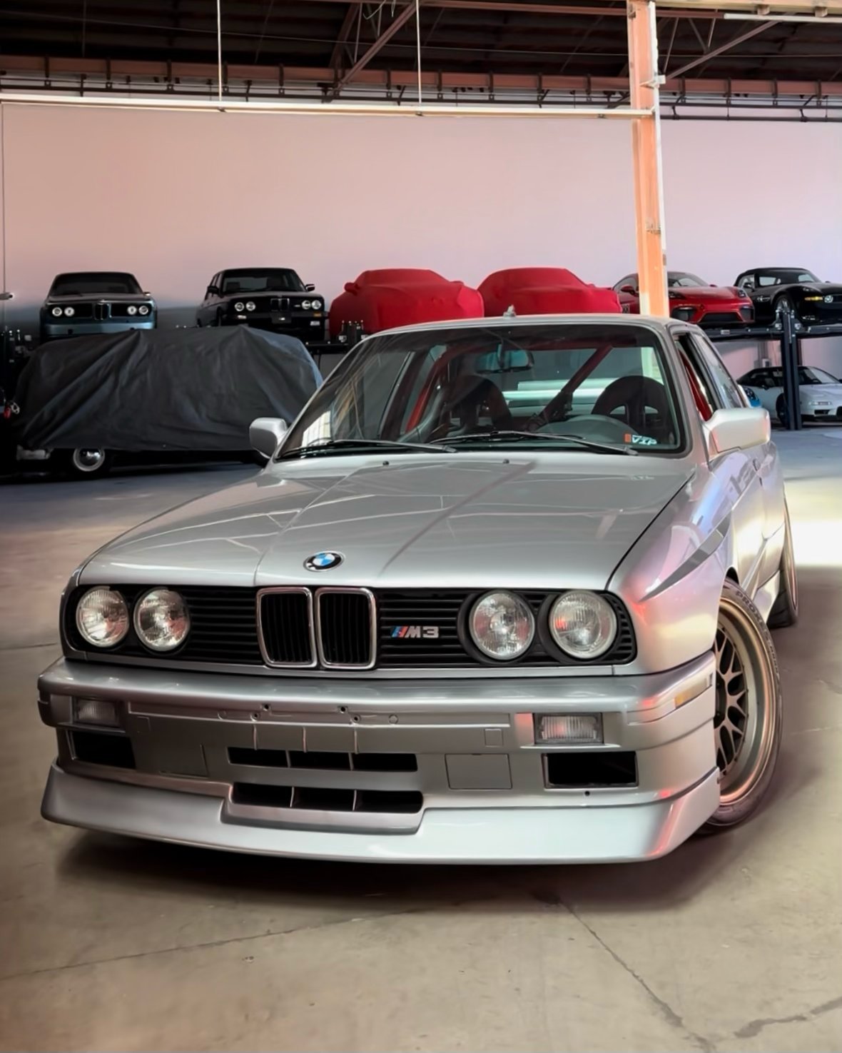 Silver BMW M3 sports car in warehouse with covered classic cars in background