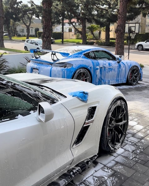 Two sports cars being washed in a residential driveway - white car in foreground and blue car in background on a tree-lined street