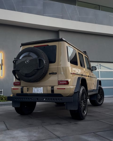 Beige Mercedes-Benz G-Class SUV with spare tire on back, parked in front of modern building with gray facade
