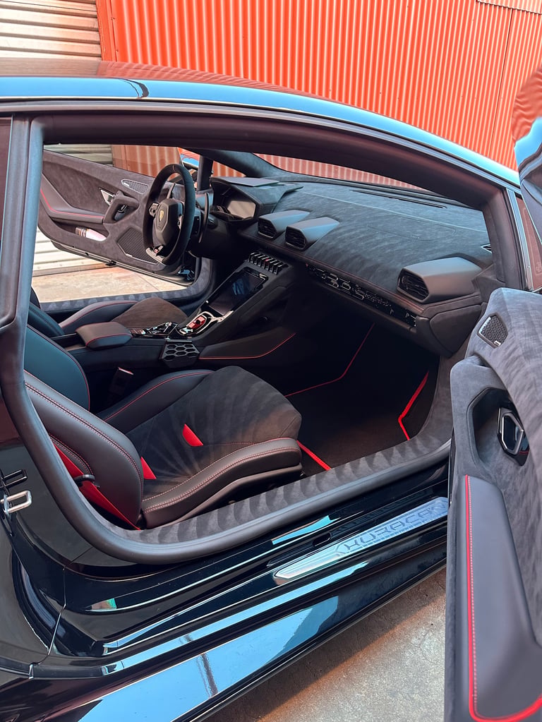 Interior view of a sports car with red and black racing seats, carbon fiber dashboard, and open door against a corrugated metal building backdrop