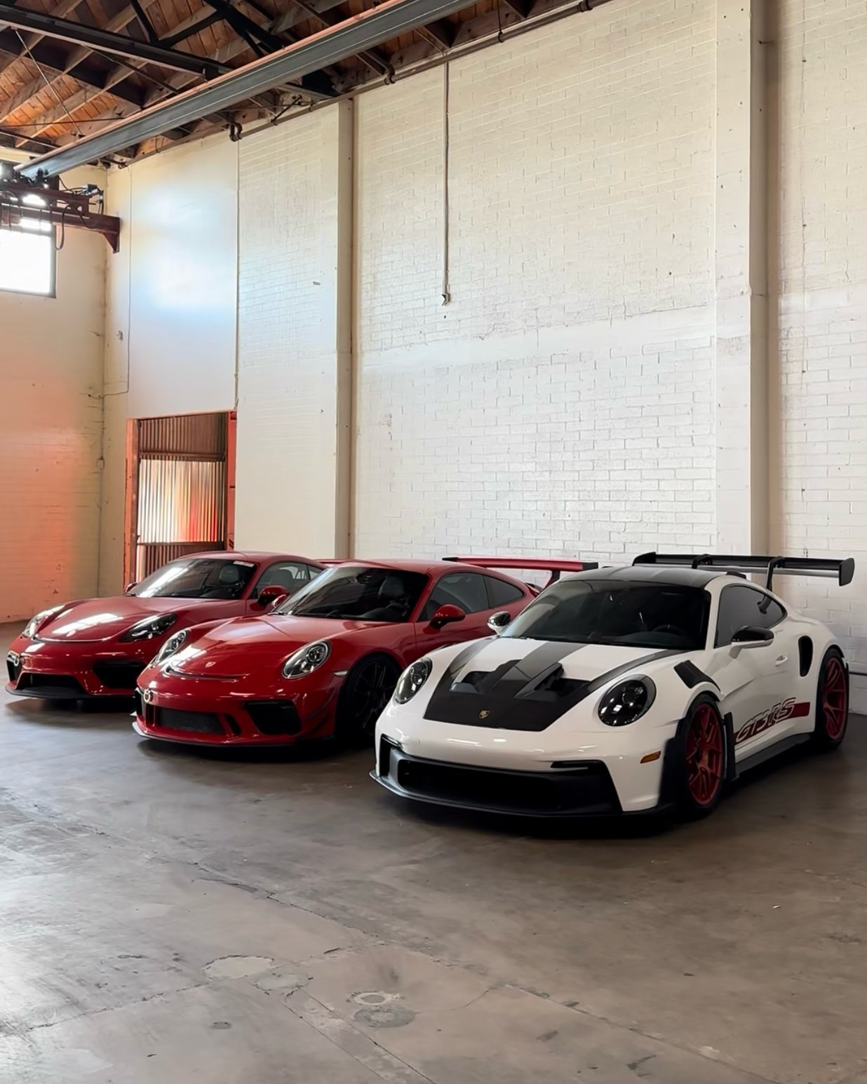 Three sports cars parked in an industrial warehouse: two red Ferraris and one white Porsche with black accents