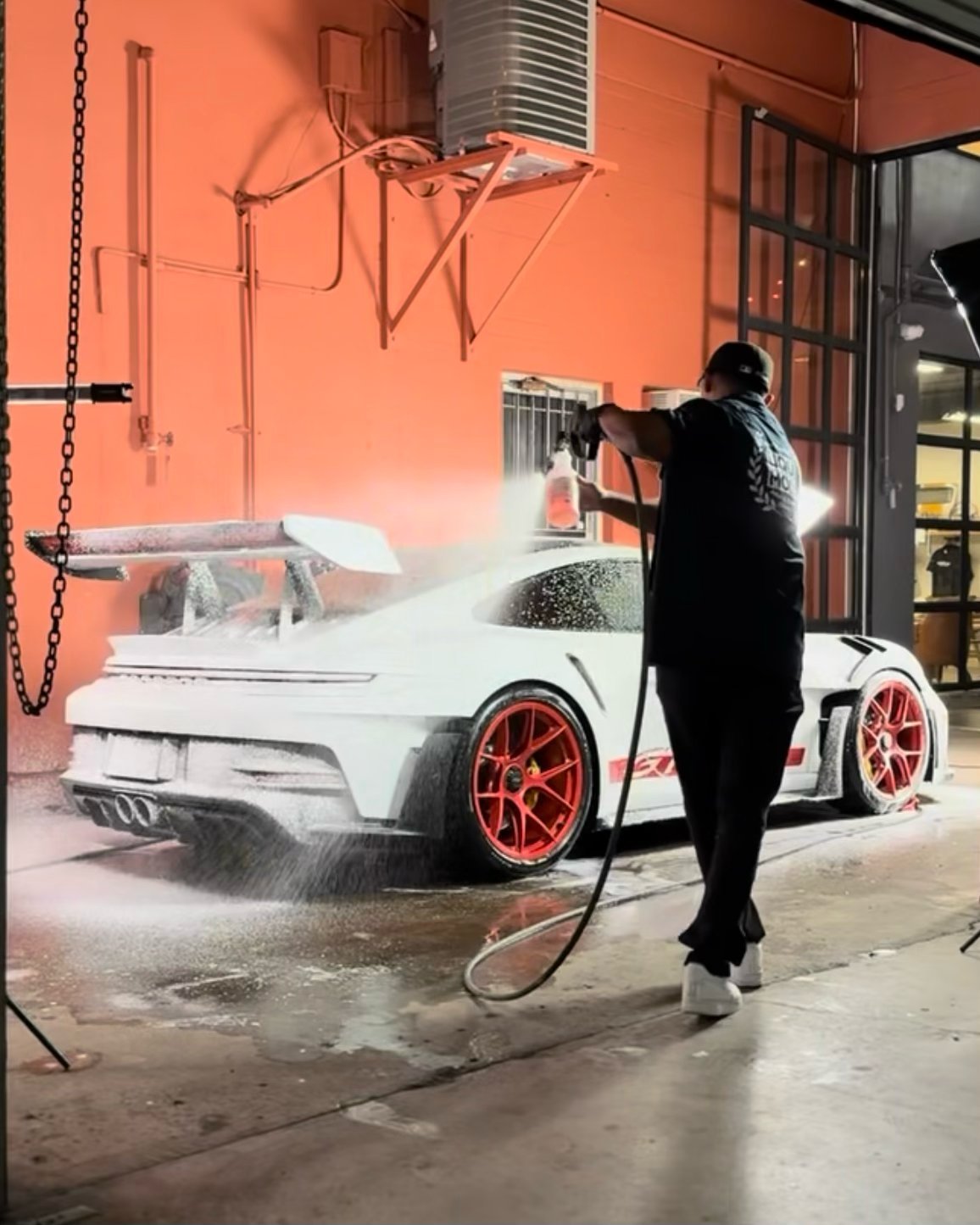 Person washing a white sports car with red wheels using a pressure washer in an orange-walled garage