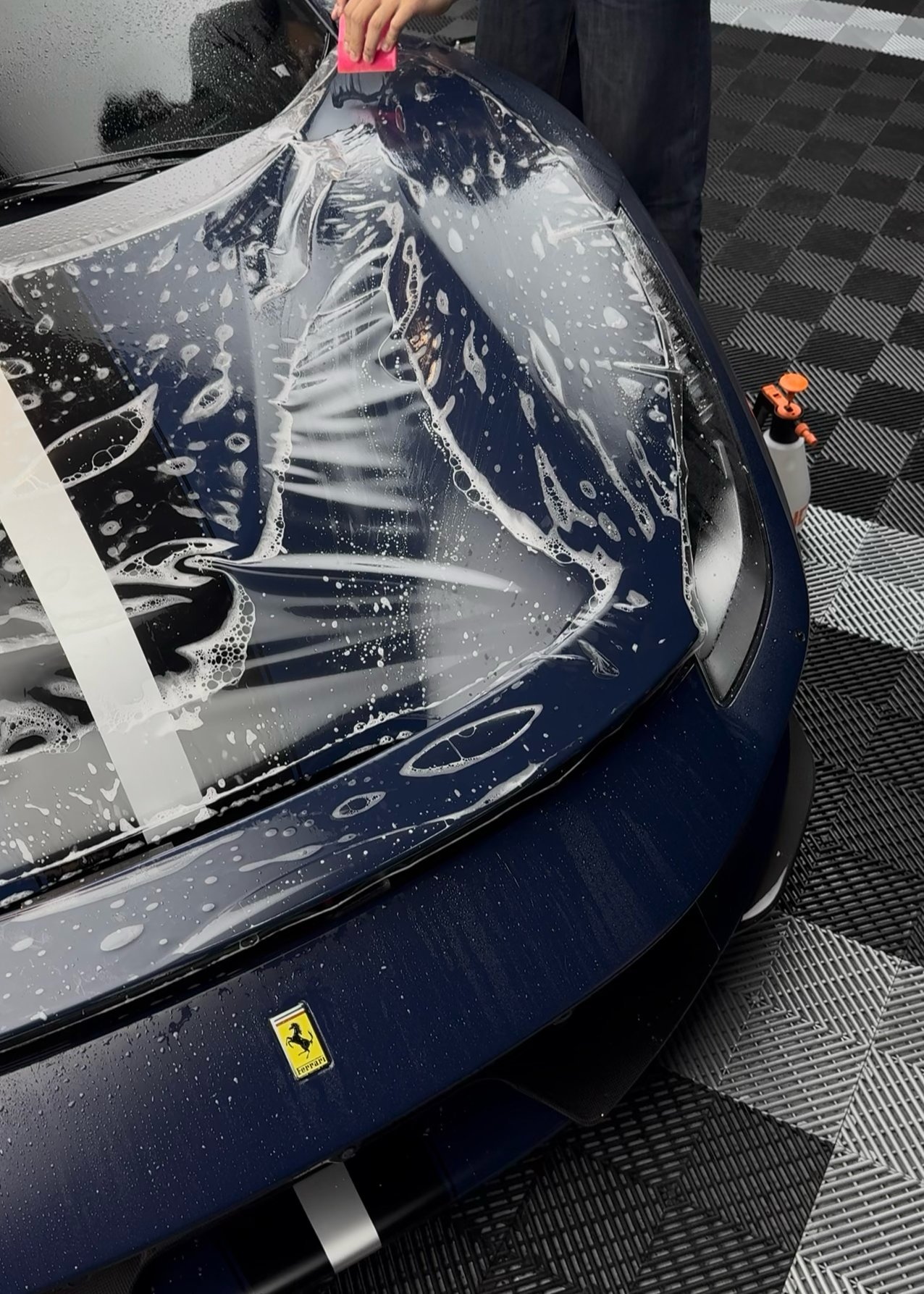 Person removing protective plastic wrap from a dark blue Ferrari car hood during vehicle preparation
