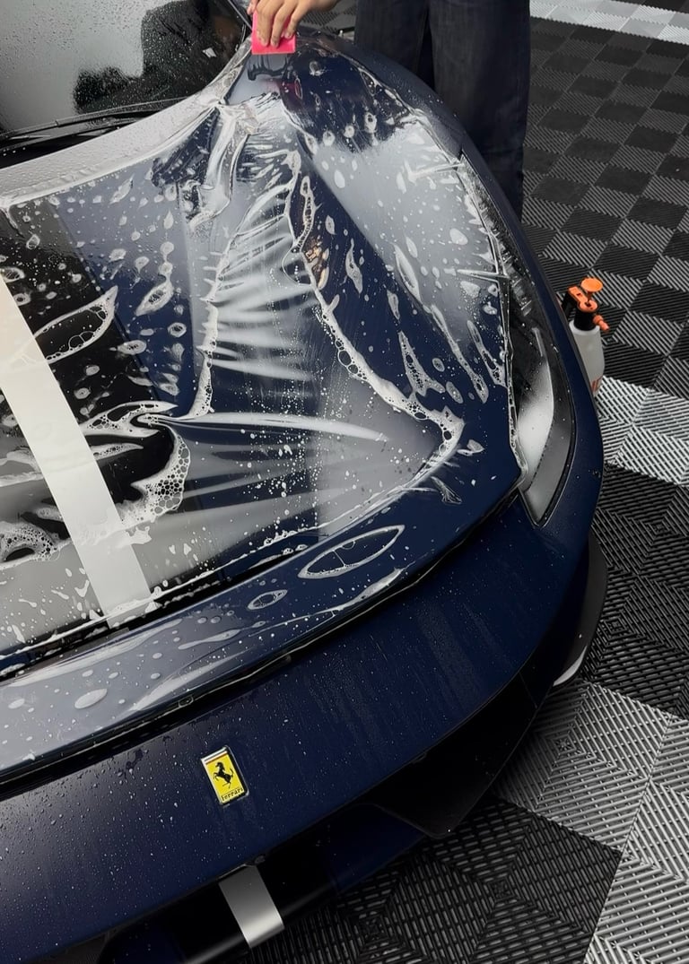 Person removing protective plastic wrap from a dark blue Ferrari car hood during vehicle preparation