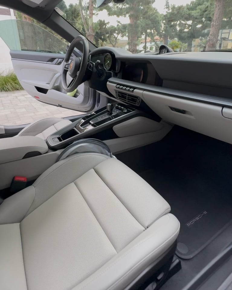 Interior view of luxury sports car with white leather seats, steering wheel, dashboard, and windshield view of trees