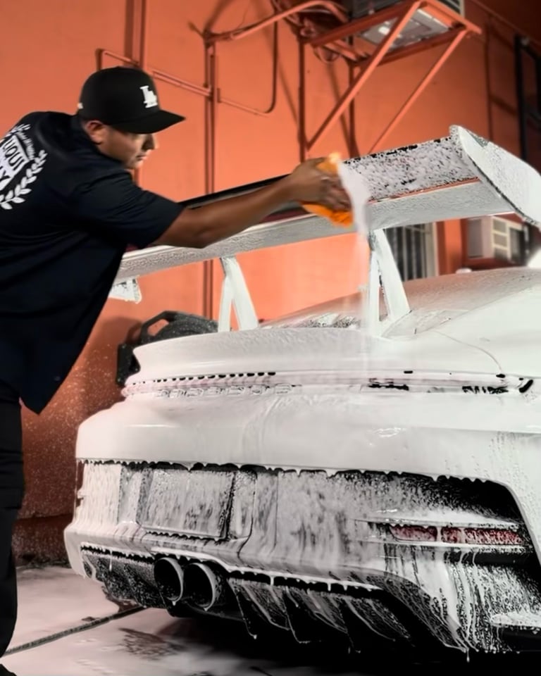 Worker in black cap and shirt washing a white car with foam and brush at an indoor car wash bay with orange wall