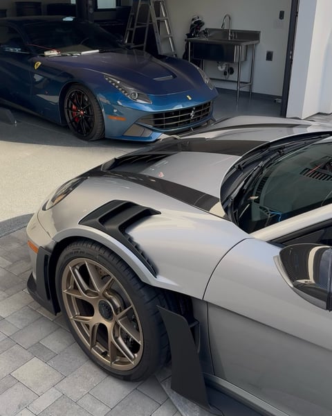 White sports car with black hood stripes and blue Ferrari in background, inside a garage or showroom
