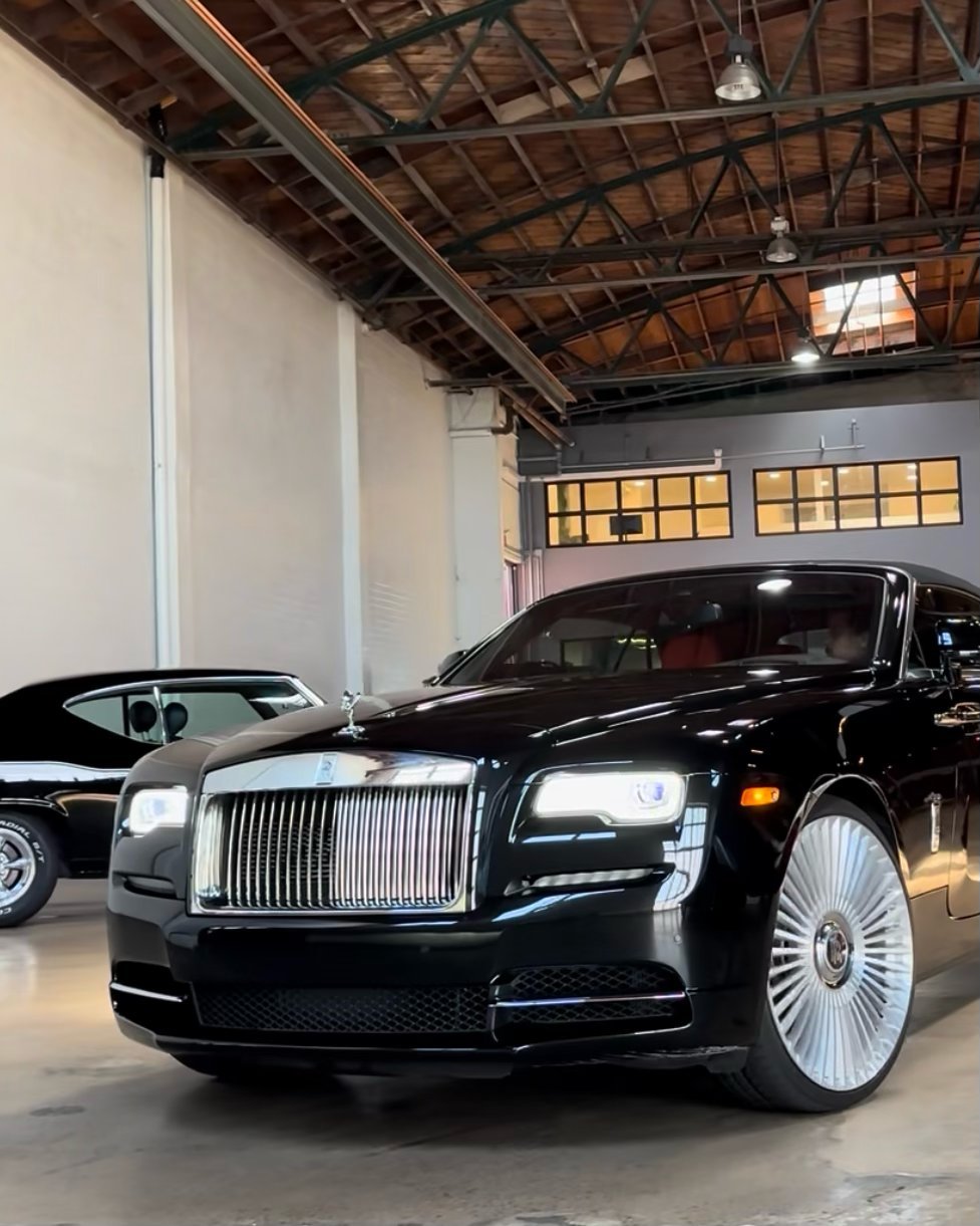 Black luxury Rolls-Royce sedan displayed in a modern garage with exposed wooden ceiling and skylights